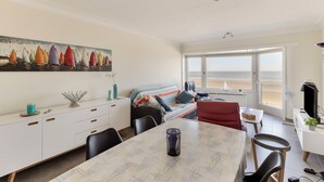 Una sala moderna con comedor, un amplio ventanal con vista a la playa y un cuadro de veleros en la pared.