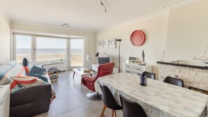 Amplia sala de estar con sofá, mesa de comedor y sillas, una silla roja, una lámpara, un reloj y vista a la playa a través de grandes ventanales.