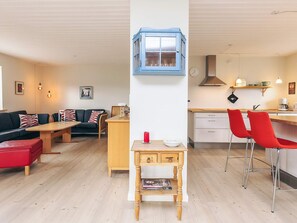 Un salon moderne avec un canapé, une table basse et un pouf rouge. On y voit une table d’appoint en bois sur laquelle sont posés un magazine et une tasse rouge. Une partie de la cuisine, avec une cuisinière, un évier et des chaises rouges, est visible.