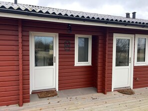 Un bâtiment en bois rouge, doté de deux portes et fenêtres blanches, d’une plaque d’immatriculation en métal et d’un paillasson.
