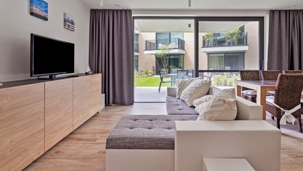 Un salon moderne doté d’un téléviseur à écran plat, d’un canapé, d’une table à manger avec des chaises, et offrant une vue sur un balcon et un espace extérieur.