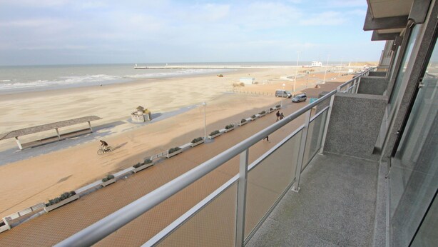 A beach view from a balcony with a railing, sandy beach, and a pier in the distance.