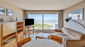 Une chambre d’hôtel dotée d’une grande fenêtre donnant sur la plage, d’un canapé, d’une table à manger et d’un téléviseur posé sur un meuble.