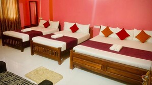 A hotel room with four single beds, each with a wooden frame and red and white bedding. There is a small table with a patterned table runner and a chair. The room has a red wall and a white floor. - Cinnamon Tree (Anuradhapura)