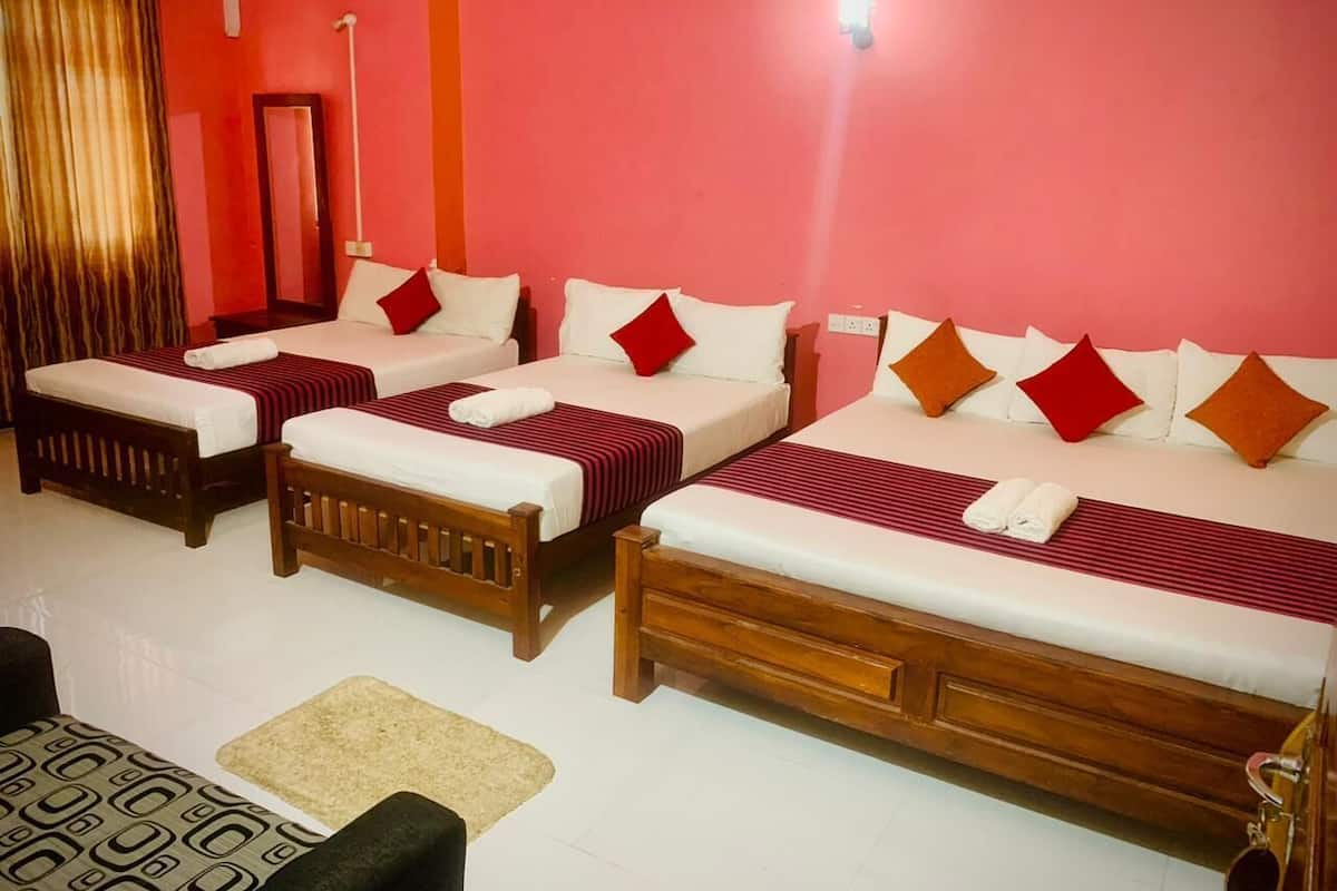 A hotel room with four single beds, each with a wooden frame and red and white bedding. There is a small table with a patterned table runner and a chair. The room has a red wall and a white floor.