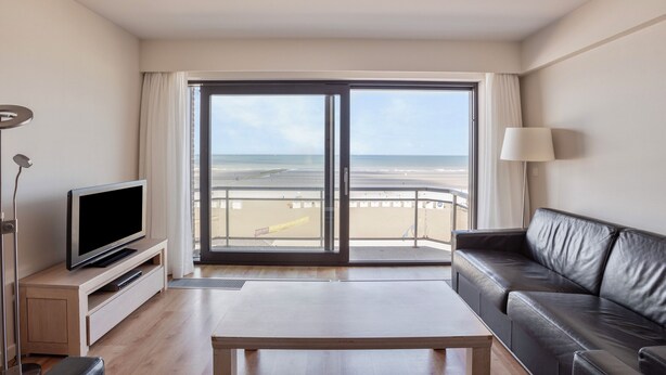 A modern living room with a television, a sofa, a coffee table, and a view of the beach through large sliding doors. - 2 bedroom accommodation in Nieuwpoort (Nieuwpoort)