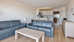 A modern living room with a grey leather sofa, a wooden coffee table, and a kitchen area in the background. - 2 bedroom accommodation in Nieuwpoort (Nieuwpoort)