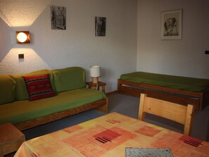 A small, cozy room with a green sofa, a wooden bed, and a table with a patterned tablecloth.