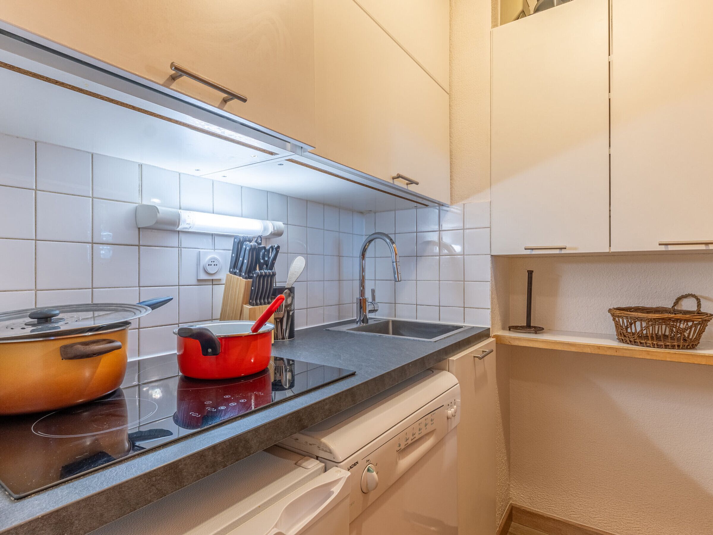 Une cuisine avec des meubles blancs, un plan de travail noir, une casserole rouge, un évier et un égouttoir à vaisselle.