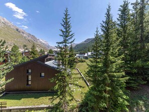 A modern wooden cabin with a sloped roof, surrounded by lush greenery and mountains in the background.