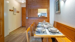 On aperçoit un coin repas avec une table en bois dressée pour quatre personnes, bordée de bancs en bois. Une cuisine avec une fenêtre et un miroir est visible au fond.