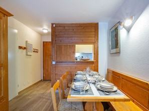 A dining area with a wooden table set for four, flanked by wooden benches. A kitchen with a window and a mirror is visible in the background.
