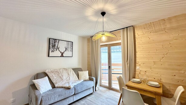 En moderne stue med en grå sofa, et træspisebord med stole, en pendellampe og et stort vindue med gardiner.