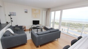A modern living room with a grey sofa, a small table, a TV, and a balcony with a view of the beach.