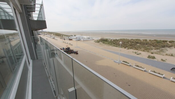 A high-rise building with a view of a beach, including a road and parked vehicles.