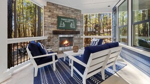 Un rincón al aire libre acogedor con chimenea de piedra, mobiliario blanco y amplias ventanas con vistas al bosque.