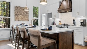 Una cocina moderna con armarios blancos, una isla grande con taburetes de bar, un refrigerador de acero inoxidable y una cafetera.