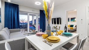 Ein moderner Essbereich mit einem weißen Tisch, gedeckt für vier Personen, umgeben von grauen Sofas. Auf dem Tisch steht eine Vase mit getrockneten Blumen, und im Hintergrund ist ein großes Fenster mit blauen Vorhängen zu sehen.