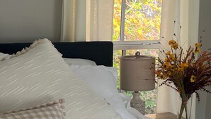 Una cama bien tendida con ropa de cama blanca y una almohada a cuadros, una lámpara de noche y un jarrón con flores sobre una mesita de noche.