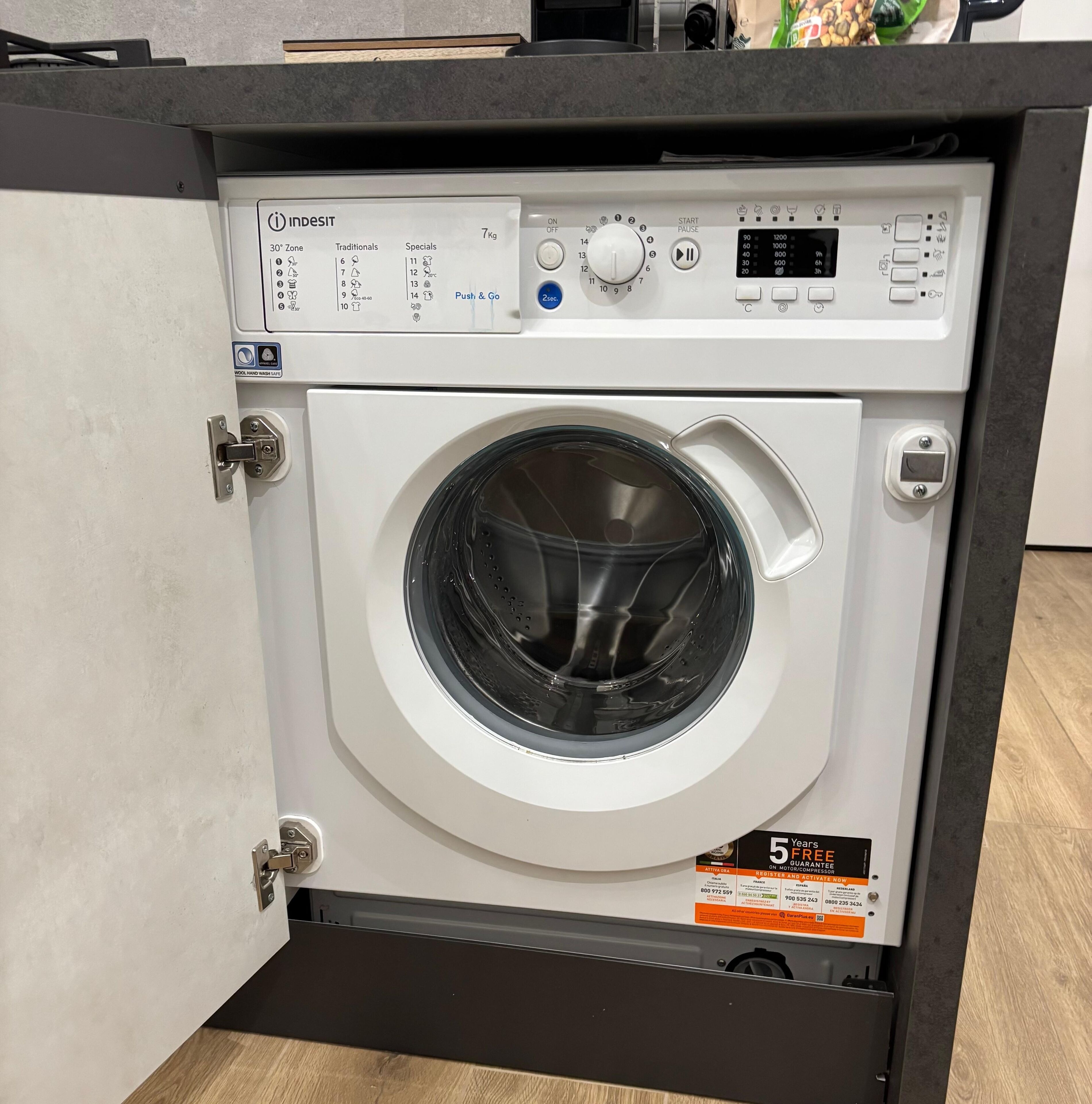 Een compacte voorlader wasmachine met een digitaal bedieningspaneel, een etiket met een capaciteit van 7 kg, en een sticker voor een gratis garantie van 5 jaar.