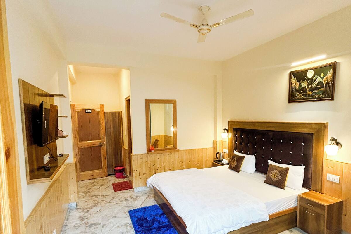 Une chambre d’hôtel avec un grand lit, une tête de lit en bois, un sol en marbre, un ventilateur de plafond, une petite table et un téléviseur fixé au mur.