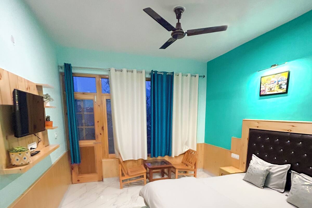 Une chambre avec un lit, un ventilateur de plafond, une fenêtre avec des rideaux, une petite table et un téléviseur fixé au mur.