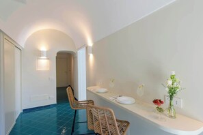 A modern interior with a blue-tiled floor, a hallway leading to another room, wicker chairs, a white counter with a vase of flowers, and wall-mounted lighting.