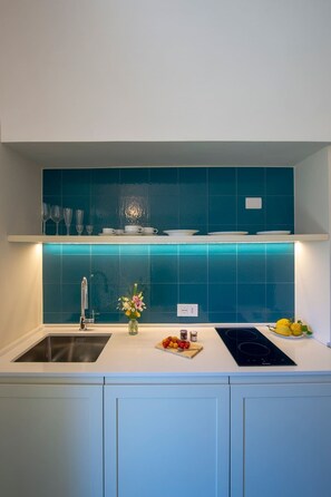 A modern kitchen with a white countertop, a stainless steel sink, a black induction cooktop, and teal backsplash tiles.