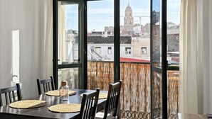 Un coin repas avec une table dressée pour quatre personnes, de grandes fenêtres offrant une vue sur des immeubles et une tour, et un rideau en tissu tissé.