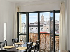Comedor con una mesa dispuesta para cuatro, ventanas amplias con vistas a edificios y una torre, y una cortina tejida.