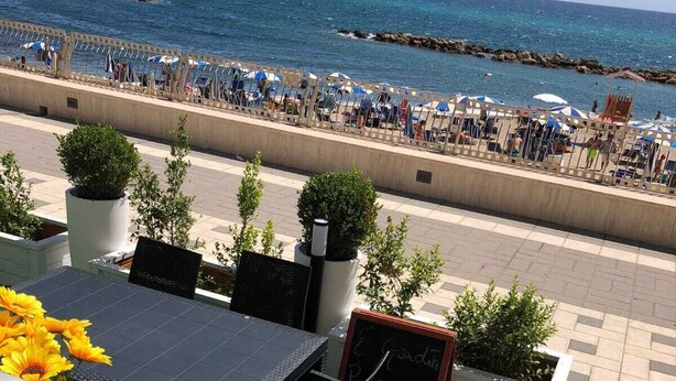 A terrace with a table, chairs, and a pizza on a stand. There are potted plants and a view of the beach with people in the background. - Summer events agr pt (Torre Melissa)