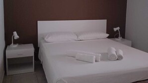 A neatly made bed with white linens and towels, a bedside table with a lamp, and a plain wall. - Summer events agr pt (Torre Melissa)