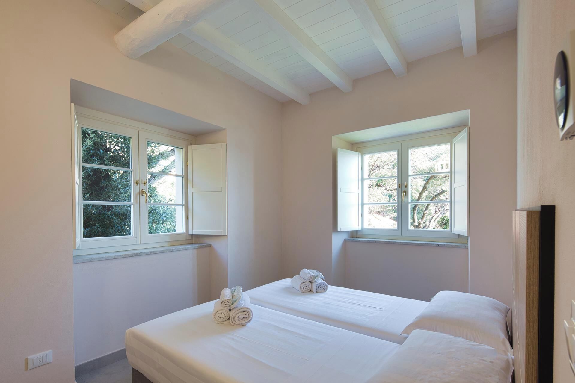 Une chambre avec deux lits, de grandes fenêtres avec des volets blancs et un plafond avec des poutres apparentes.