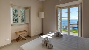 A bedroom with a bed, a wooden bench, a lamp, and a view of the sea through a large window. - Campetti (Genoa)