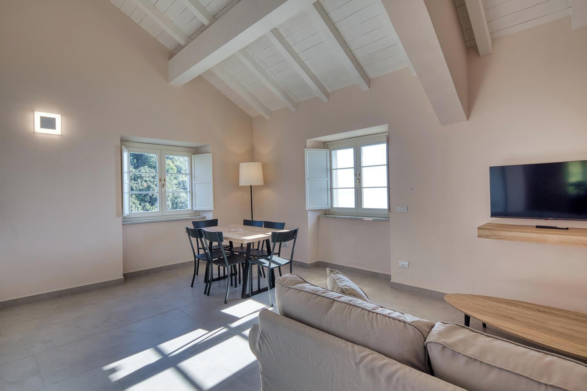 Un salón amplio con techo alto, una mesa de comedor con sillas, un sofá, un televisor montado en la pared y ventanas que dejan entrar luz natural.