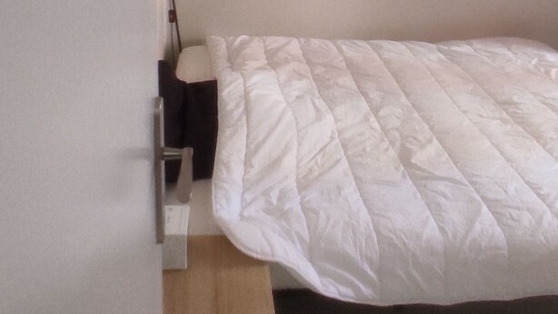 Een kleine kamer met een enkel bed, een houten nachtkastje, een lamp en een wit gebogen matras.