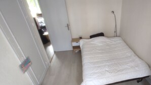 Een kleine eenpersoonskamer met een wit sprei, een nachtkastje en een wandlamp. Een deur leidt naar een gang met uitzicht op geparkeerde auto's.