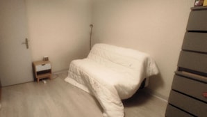Een kleine, zwak verlichte kamer met een enkel bed, een klein houten nachtkastje en een kast.