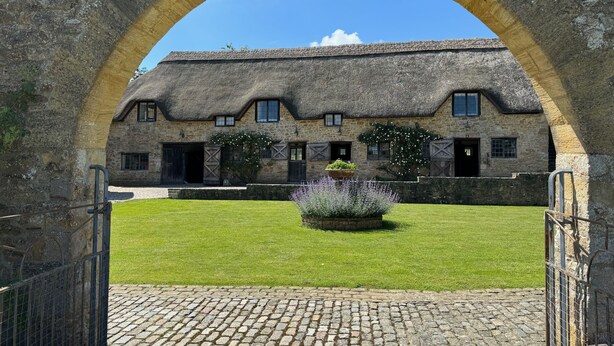 A stone building with a thatched roof, a paved courtyard, and a metal gate.