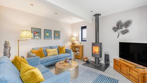 Een moderne woonkamer met een blauwe bank, gele kussens, een ronde salontafel, een open haard, een televisie en een boekenkast.
