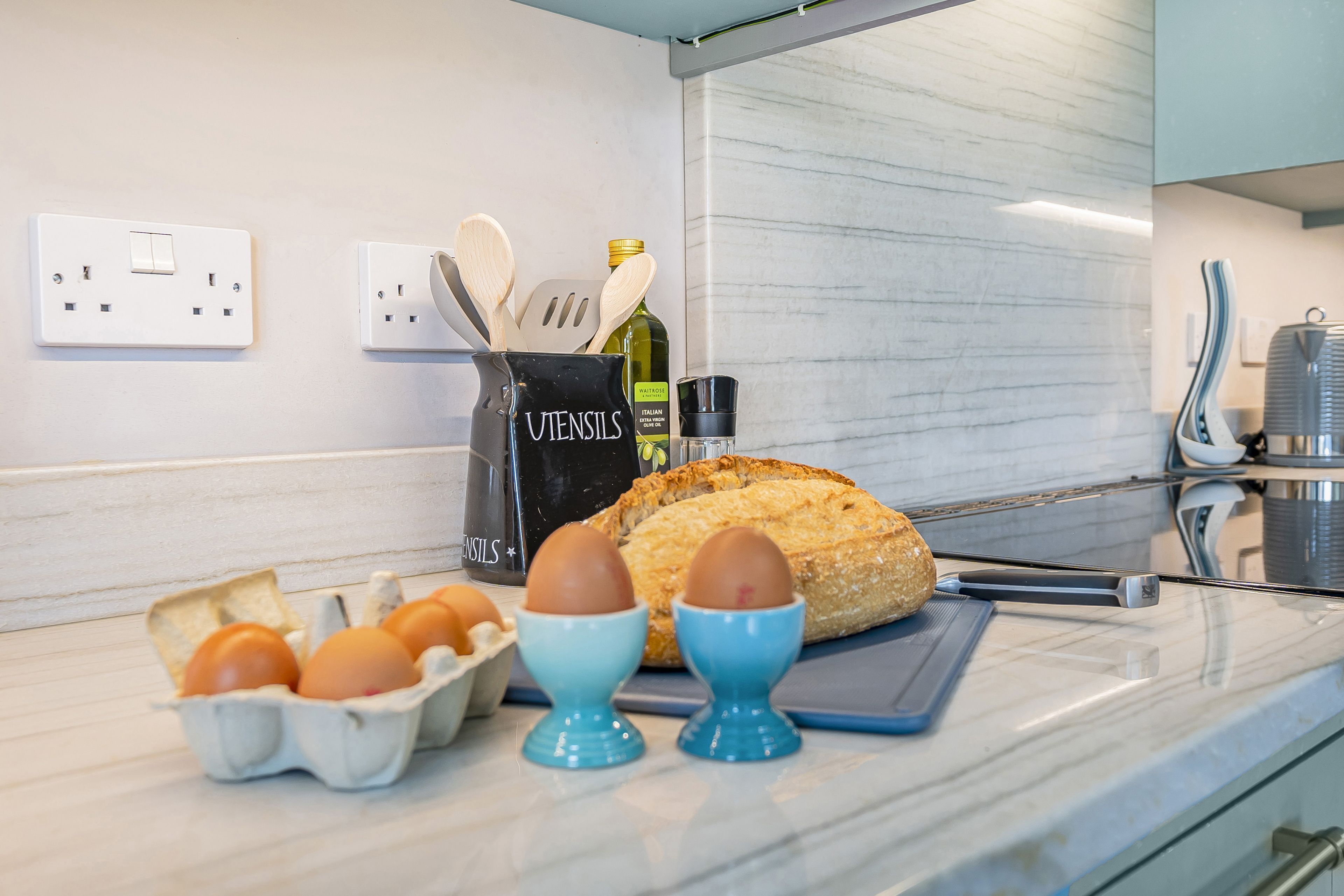 Una encimera de cocina con huevos en cajas, portavasos para huevos, una hogaza de pan y utensilios de cocina.