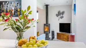 Een moderne woonkamer met een houtkachel, een televisie en een eethoek met een schaal fruit en bloemen.
