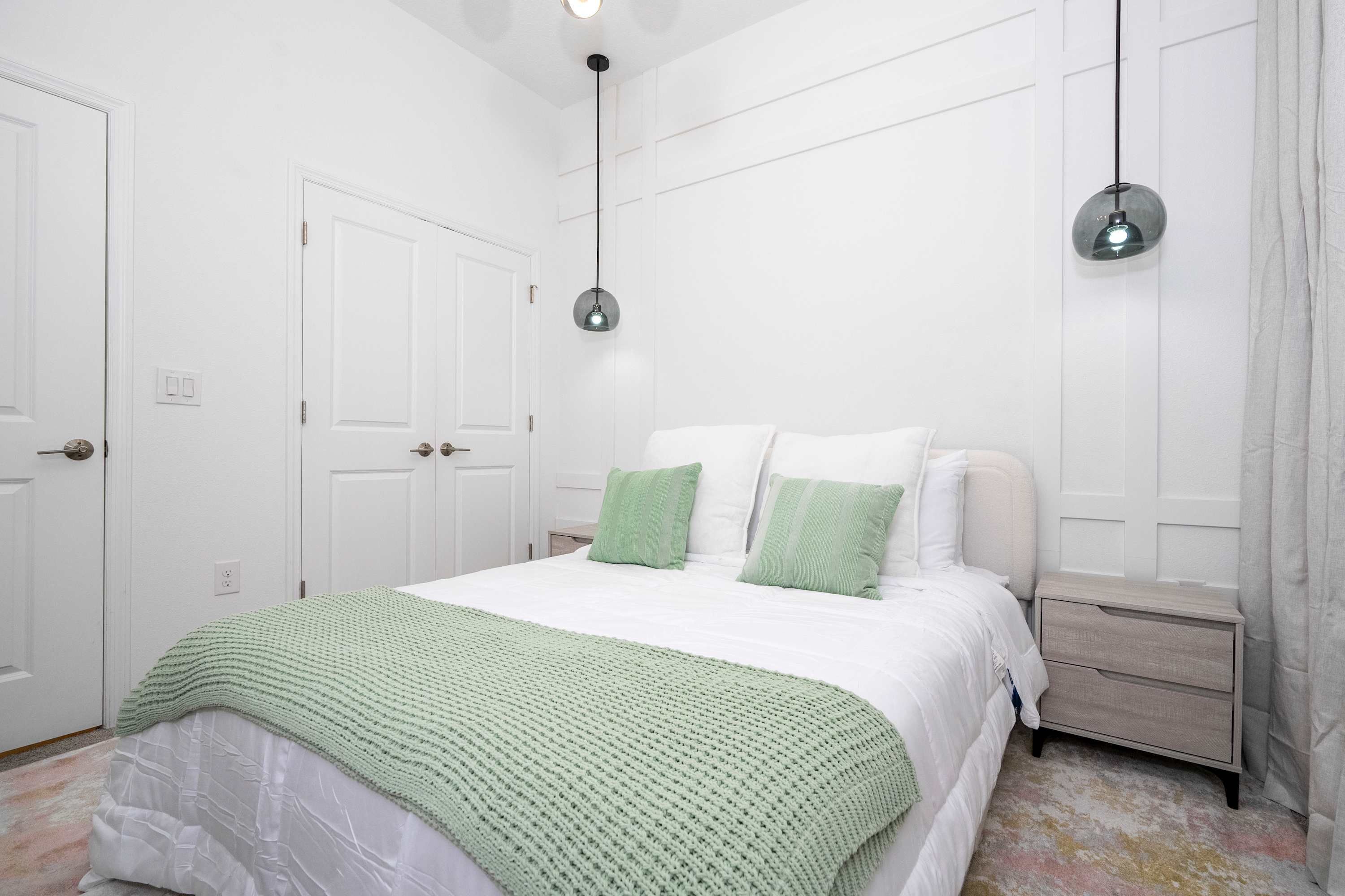 A bedroom with a bed, green and white pillows, a nightstand, and two pendant lights.