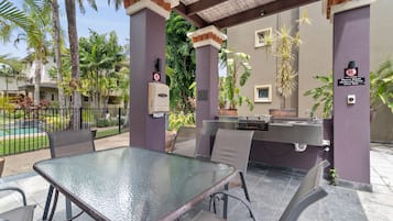 An outdoor patio with a glass table and chairs, a barbecue grill, and a pool in the background.