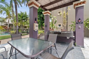 Een terras buiten met een glazen tafel en stoelen, een barbecue en een zwembad op de achtergrond.