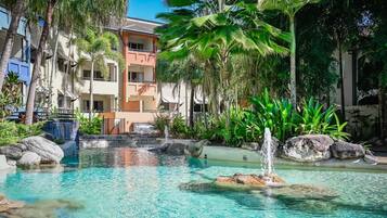 A pool with clear water, surrounded by rocks and palm trees, with a building in the background.