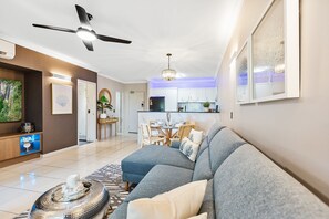 Una sala moderna con un sofá gris, un comedor con mesa y sillas, una cocina con armarios blancos y un ventilador de techo.