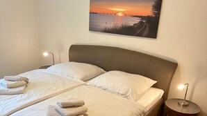 A double bed with white linens and towels, a round nightstand with a lamp, and a sunset painting on the wall. - Coastal Comfort With Garden (Fehmarn)