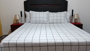 A neatly made bed with a quilted white and black pattern, flanked by two bedside tables with lamps, a large wall painting, and a geometric patterned rug. - Hostal Deluxe Py offers you comfort, friendliness and (Asunción)