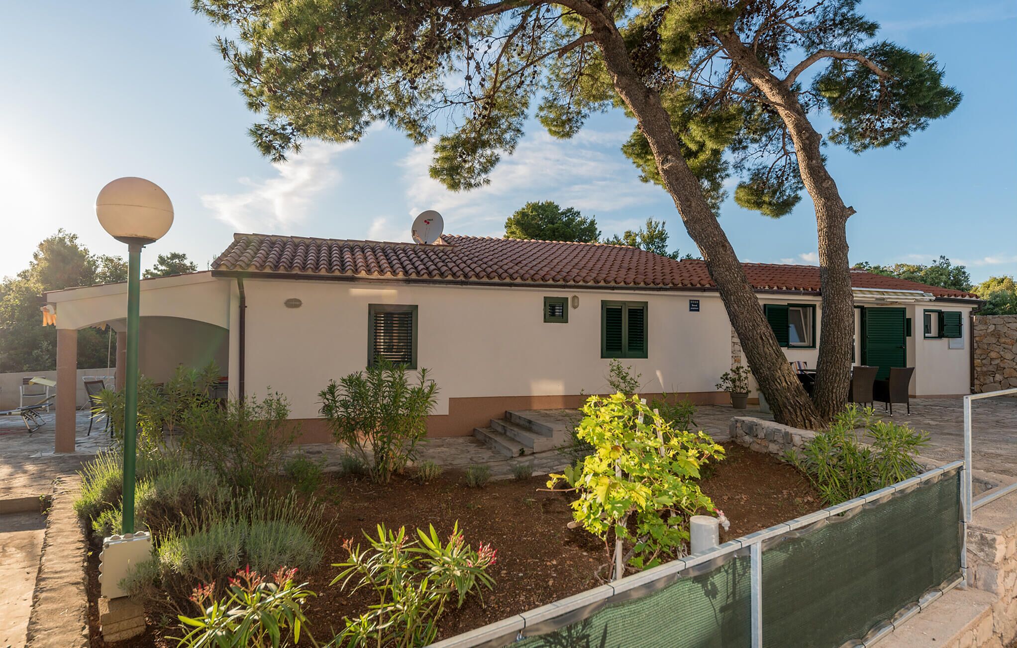 A two-story house with a tiled roof, green shutters, and a small garden with various plants and a tree.